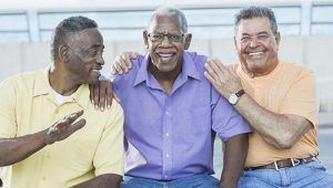 A group of three multiracial senior men sitting together on bench outdoors. The Caucasian man, in his 80s, has his hands on the shoulders of the African American man sitting in the middle. They are smiling at the camera.