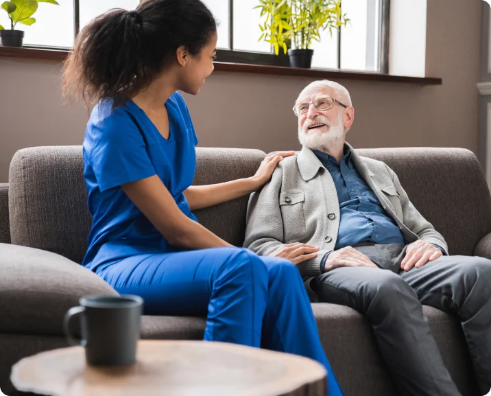 A nurse engages in conversation with an elderly man seated on a couch, highlighting compassionate senior care.