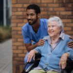 Elderly woman in a wheelchair smiling outdoors with a caring male nurse standing behind her