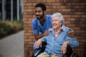 Elderly woman in a wheelchair smiling outdoors with a caring male nurse standing behind her