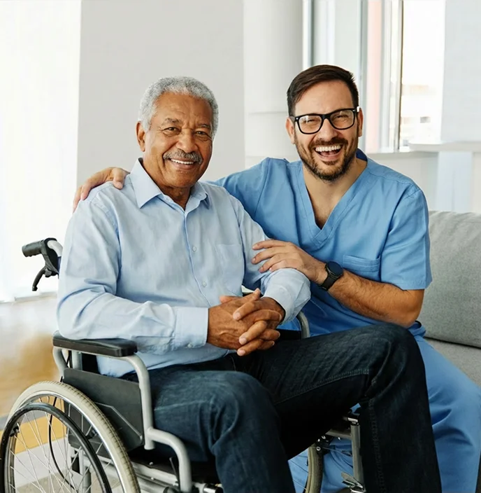 Professional caregiver sitting beside smiling senior man in wheelchair during home care visit