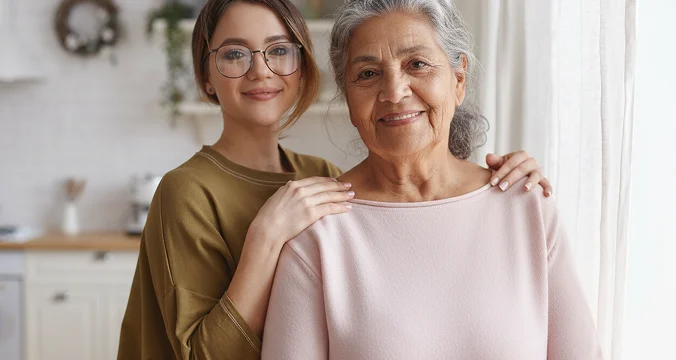 Young caregiver standing beside smiling senior woman at home