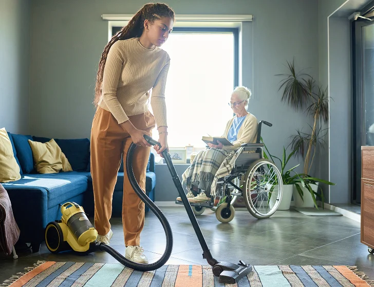Home caregiver vacuuming living room while senior woman relaxes in wheelchair nearby
