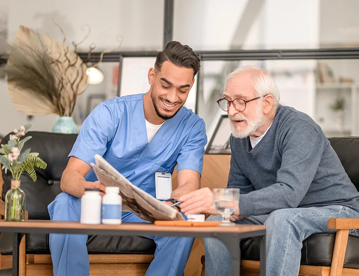 Friendly caregiver reading newspaper together with senior man during home visit