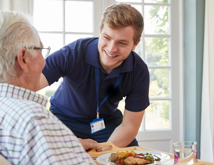 Young caregiver serving healthy meal to senior man at home