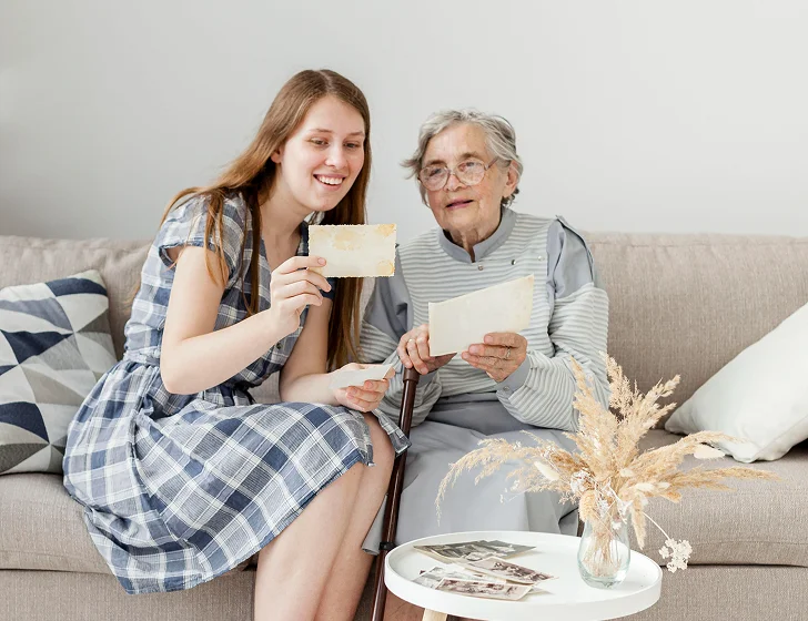 Young caregiver and elderly woman looking through family photos and memory keepsakes together